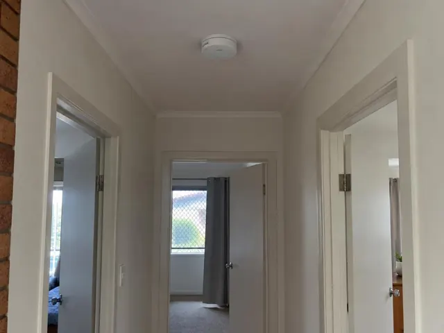 Ceiling smoke alarm in a narrow hallway with doorways and brick wall detail in Pimpama